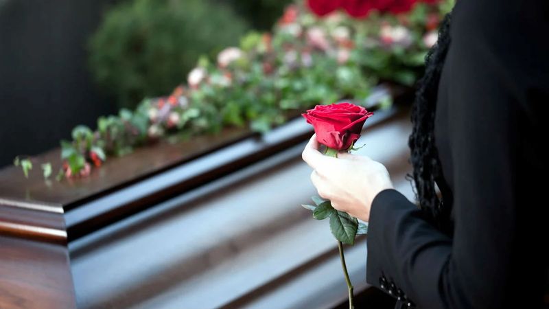 Eine Frau in schwarzer Kleidung, die eine rote Rose in der Hand hält, während sie vor einem Holzsarg steht, der mit Blumen geschmückt ist.