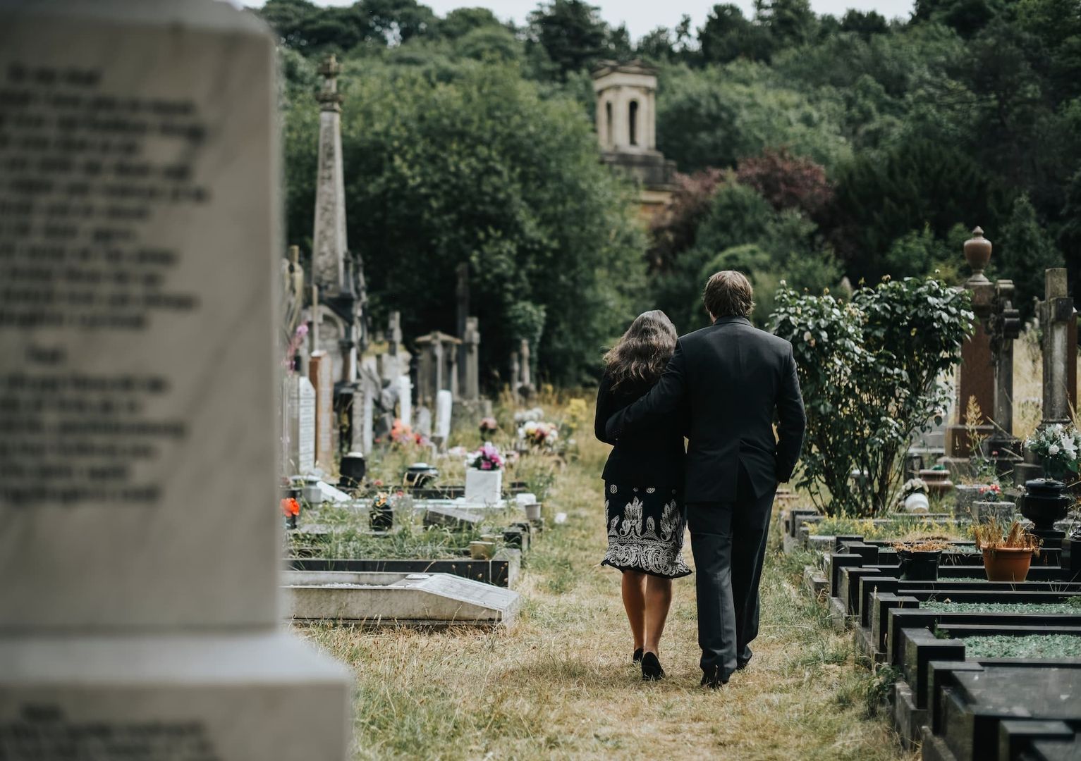 Ein Paar in Trauerkleidung, von hinten gesehen, geht durch einen Friedhof. Sie halten sich gegenseitig fest.